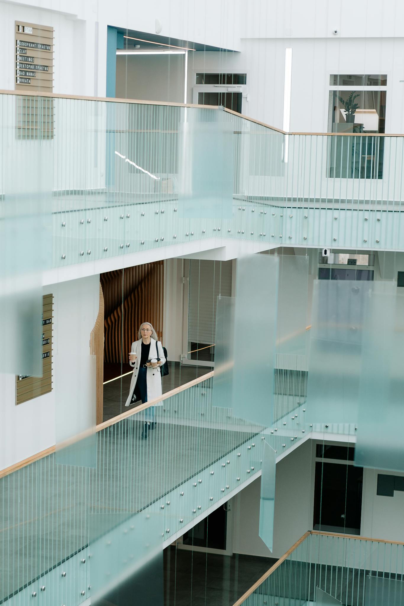 Woman Walking Through A Bright Modern Office Corridor With Glass Railings Holding Coffee And Documents. 5971046 1366x2048
